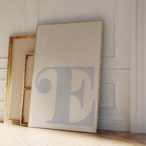 May include: Two canvases leaning against a white wall. The larger canvas is white with a light blue letter "E" in the center. The smaller canvas is brown with a wooden frame.