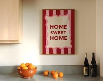 Puede incluir: Una impresión enmarcada con rayas rosas y rojas y un cuadrado blanco en el centro. El texto "HOME SWEET HOME" está escrito en letras rojas dentro del cuadrado blanco. Hay naranjas en un tazón marrón y dos botellas de vino en una encimera gris.