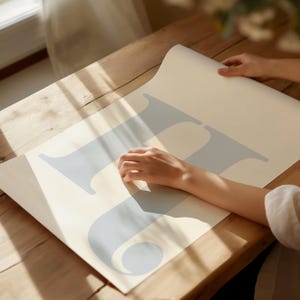 May include: A light blue letter "H" printed on a white sheet of paper. The paper is rolled up and a person's hand is touching the paper.