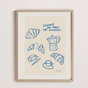 Op de afbeelding: Een ingelijst print met een licht beige achtergrond en blauwe illustraties van croissants, een koffiepot, boter, een mes en een kopje koffie. De tekst "Sundays are made for croissants" is geschreven in blauwe cursieve letters.