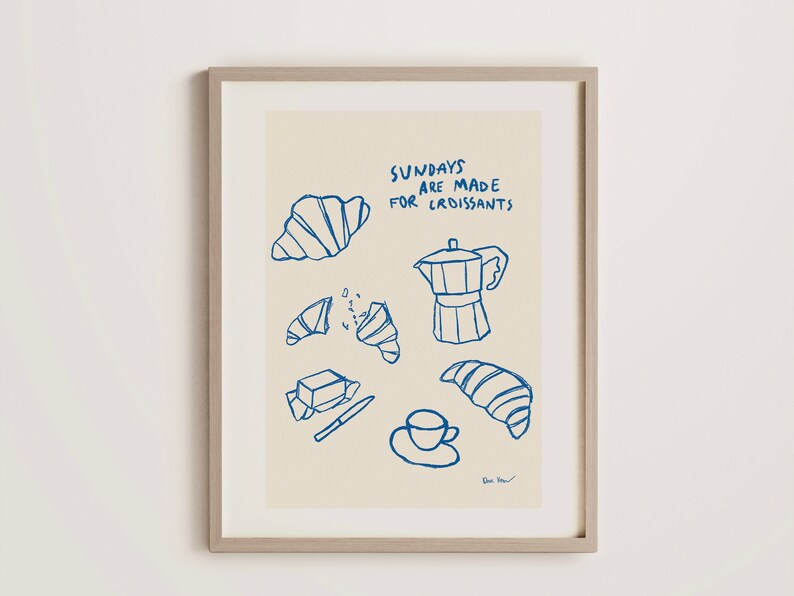 Op de afbeelding: Een ingelijst print met een licht beige achtergrond en blauwe illustraties van croissants, een koffiepot, boter, een mes en een kopje koffie. De tekst "Sundays are made for croissants" is geschreven in blauwe cursieve letters.
