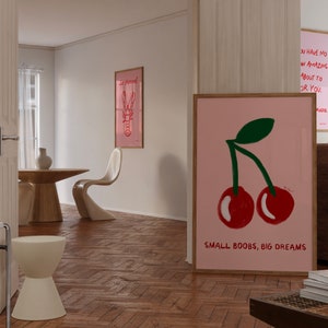 K&ouml;nnte beinhalten: Ein rosa-wei&szlig; gerahmtes Poster mit einer Zeichnung von zwei roten Kirschen und dem Text "SMALL BOOBS, BIG DREAMS". Das Poster h&auml;ngt an einer wei&szlig;en Wand in einem Raum mit Holzboden.