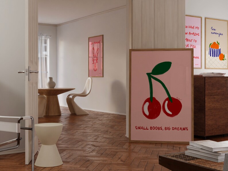 K&ouml;nnte beinhalten: Ein rosa-wei&szlig; gerahmtes Poster mit einer Zeichnung von zwei roten Kirschen und dem Text "SMALL BOOBS, BIG DREAMS". Das Poster h&auml;ngt an einer wei&szlig;en Wand in einem Raum mit Holzboden.