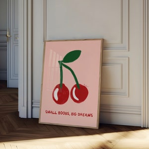 K&ouml;nnte beinhalten: Ein rosa Poster mit einer Grafik von zwei roten Kirschen mit gr&uuml;nen Stielen und Bl&auml;ttern. Der Text "SMALL BOOBS, BIG DREAMS" ist unter den Kirschen gedruckt.