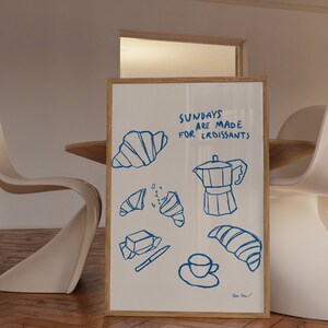 Op de afbeelding: Een wit ingelijst print met blauwe inkttekeningen van croissants, een koffiepot, een kopje koffie en een boterstaaf. De tekst op de print luidt "Zondagen zijn gemaakt voor croissants".