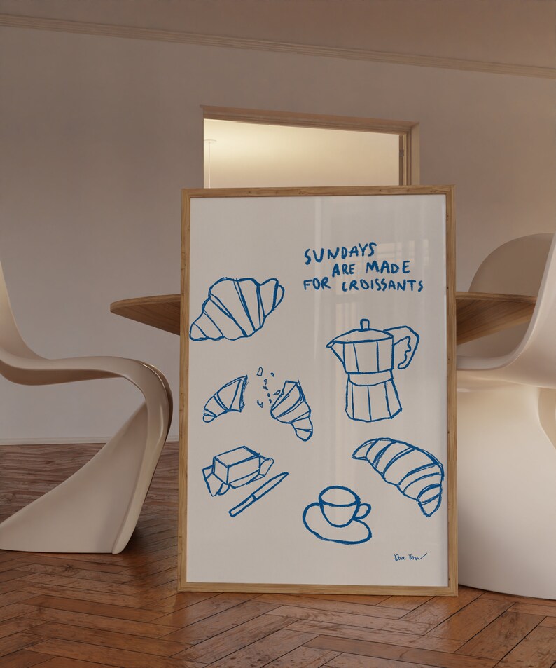 Op de afbeelding: Een wit ingelijst print met blauwe inkttekeningen van croissants, een koffiepot, een kopje koffie en een boterstaaf. De tekst op de print luidt "Zondagen zijn gemaakt voor croissants".