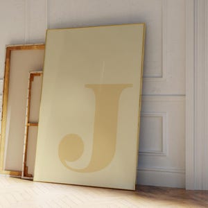 May include: A large, beige canvas leaning against a white wall, featuring a stylized letter "J" in a lighter shade of beige. A smaller canvas with a wooden frame is visible to the left. The room has a light, airy feel.