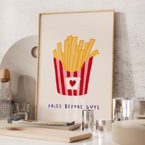 May include: A framed art print featuring a cartoon illustration of a container of golden french fries. The container is red and white striped with a heart. The text "FRIES BEFORE GUYS" is below the fries.