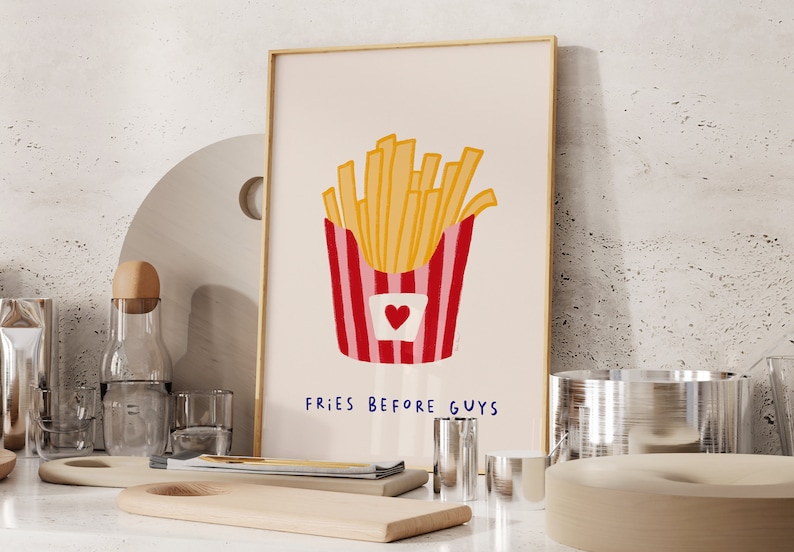 May include: A framed art print featuring a cartoon illustration of a container of golden french fries. The container is red and white striped with a heart. The text "FRIES BEFORE GUYS" is below the fries.