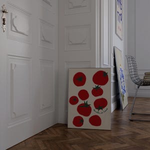 May include: A framed art print featuring a collection of red tomatoes with green stems on a beige background. The print is leaning against a white wall in a room with a wooden floor. Additional art pieces and a metal chair are visible.