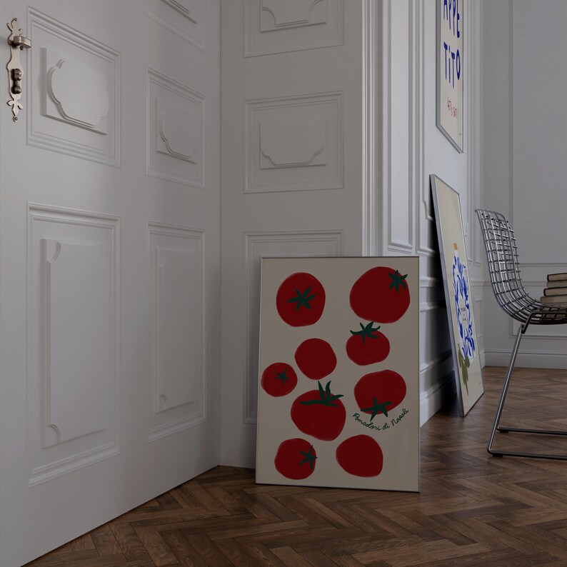 May include: A framed art print featuring a collection of red tomatoes with green stems on a beige background. The print is leaning against a white wall in a room with a wooden floor. Additional art pieces and a metal chair are visible.