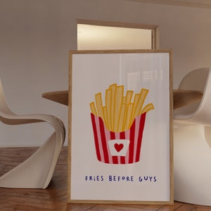 May include: A framed art print featuring a colorful illustration of a container of french fries. The fries are yellow, and the container is red and pink with a heart. The text "FRIES BEFORE GUYS" is written in blue.