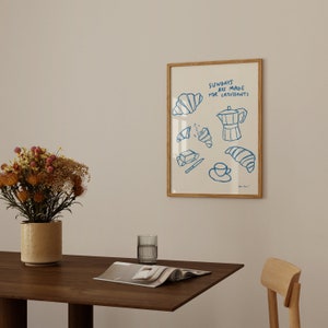 Op de afbeelding: Een ingelijst print met een blauwe illustratie van croissants, boter, een koffiepot en een kopje koffie. De tekst op de print luidt "Zondagen zijn gemaakt voor croissants".