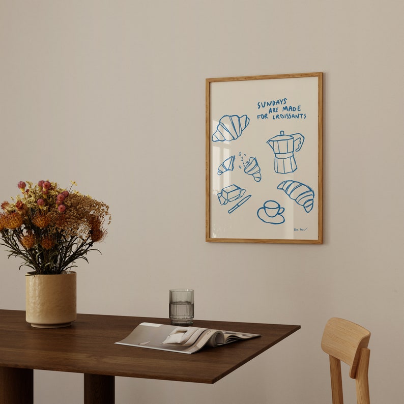 Op de afbeelding: Een ingelijst print met een blauwe illustratie van croissants, boter, een koffiepot en een kopje koffie. De tekst op de print luidt "Zondagen zijn gemaakt voor croissants".