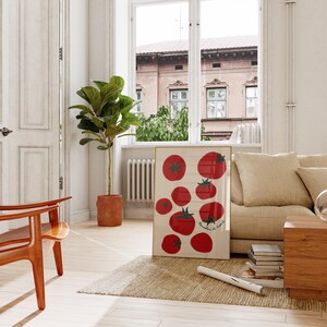 May include: A framed art print featuring a collection of red tomatoes with green stems on a cream background. The print is displayed in a living room setting with a beige sofa, wooden chair, and a potted plant. The text "Pomodori di Puglia" is visible.