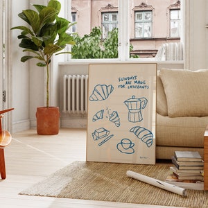 Op de afbeelding: Een ingelijste illustratie met de tekst "Sundays are made for croissants" in blauwe letters. De illustratie toont een koffiepot, croissants en een mes op een witte achtergrond.