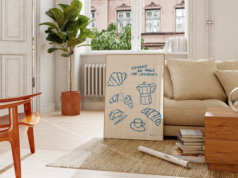 Op de afbeelding: Een ingelijste illustratie met de tekst "Sundays are made for croissants" in blauwe letters. De illustratie toont een koffiepot, croissants en een mes op een witte achtergrond.