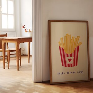 May include: A framed print featuring a cartoon illustration of a container of french fries. The container is red and white striped with a heart. The text "FRIES BEFORE GUYS" is below the fries. The frame is brown.