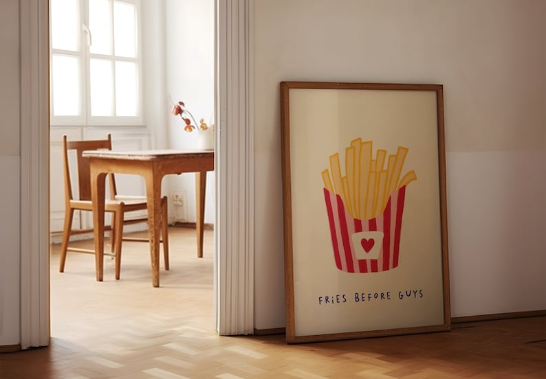 May include: A framed print featuring a cartoon illustration of a container of french fries. The container is red and white striped with a heart. The text "FRIES BEFORE GUYS" is below the fries. The frame is brown.