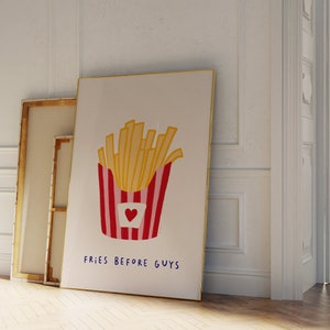 May include: A framed art print featuring a cartoon illustration of a container of french fries. The fries are yellow, and the container is red and white striped with a heart. The text "FRIES BEFORE GUYS" is written below.