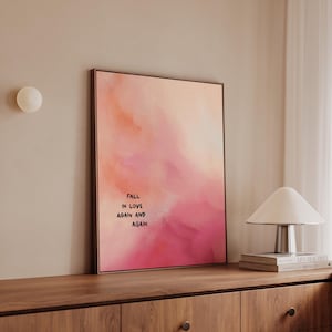 Könnte beinhalten: Gerahmter Kunstdruck mit einem Aquarell-Hintergrund in Rosa- und Pfirsichtönen. Der Text "FALL IN LOVE AGAIN AND AGAIN" ist zentriert. Der Druck wird auf einem Holzschrank präsentiert.