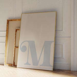 Könnte beinhalten: Drei gerahmte Leinwände lehnen an einer weißen Wand und einem Holzboden. Die größte Leinwand ist hellbeige mit einem großen, hellblauen Buchstaben "M" bedruckt. Die anderen beiden Leinwände haben goldene Rahmen.