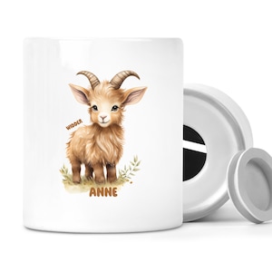 Peut inclure: Tirelire en céramique blanche avec une illustration à l'aquarelle d'un chevreau. Le texte "WIDDER" et "ANNE" est visible. Le chevreau est brun avec de grandes oreilles et des cornes incurvées, debout sur de l'herbe verte.