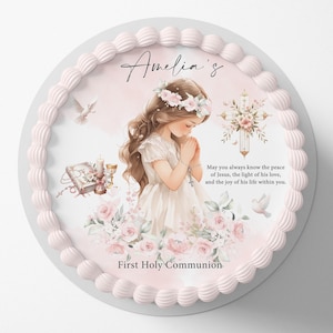Pode incluir: Um bolo redondo com cobertura rosa e uma borda decorativa. O bolo apresenta uma ilustração de uma menina com um vestido branco, uma cruz, pombas e os textos "Amelia's" e "Primeira Comunhão".