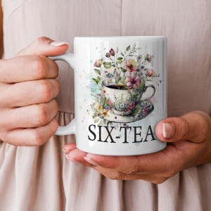 May include: White ceramic mug with a floral teacup design and the text "SIX-TEA". The teacup and saucer are surrounded by pink, yellow, and purple flowers. The mug is held in a person's hands.