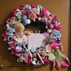 May include: A pink and white Christmas wreath with a gingerbread person, candy canes, and a sign that says "Mrs. Claus Baker".