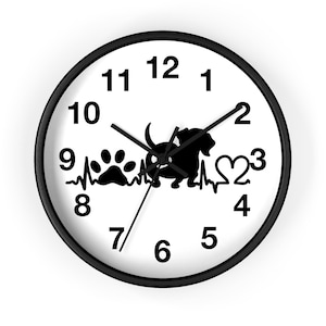 May include: A round wall clock with a black frame and a white face. The center features a black silhouette graphic of a dog, a paw print, and a heart, all connected by a horizontal heartbeat line. The clock has black hour, minute, and second hands.