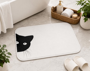 Peut inclure: Un tapis de bain rectangulaire blanc orné d'un motif de tête de chat noir sur le côté gauche. Le design du chat comprend deux yeux blancs et de fines moustaches noires. Le tapis présente des coins arrondis et une texture douce et moelleuse.