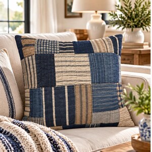 May include: A square throw pillow featuring a patchwork design of various blue, navy, and beige fabric squares. The pattern includes stripes, checks, and solid textures with visible quilted stitching throughout the surface of the pillow.