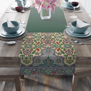 May include: A table setting with a decorative runner featuring an intricate floral pattern in beige, red, green, and blue. The table is set with teal-colored bowls, plates, wine glasses, and silverware. A vase of pink lilies adds a touch of color.