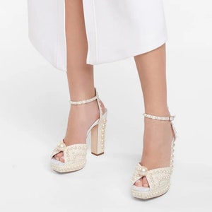 Puede incluir: Sandalias blancas de plataforma con un diseño adornado con perlas. Las sandalias tienen un tacón grueso de bloque y una correa de tobillo con cierre de hebilla.