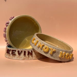 May include: Three ceramic pet bowls in a stack. The largest bowl is light yellow with a pink rim. The base of the bowl reads "KEVIN" in brown letters. The middle bowl reads "CINCY HOF" in yellow letters.