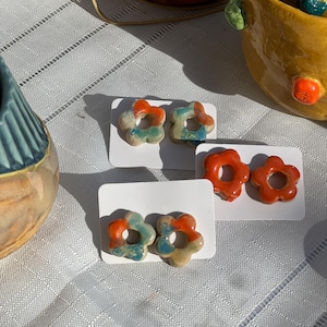 Op de afbeelding: Drie paar keramische bloemvormige oorstekers. Elk paar is gemonteerd op een witte kaart. De oorbellen zijn in tinten oranje, blauw en crème. De oorbellen liggen op een wit tafelkleed met andere keramische stukken op de achtergrond.