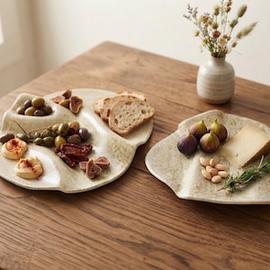 Może przedstawiać: Dwie nieregularne, kremowe ceramiczne półmiski z nakrapianą fakturą i podzielonymi sekcjami. Jeden półmisek zawiera oliwki, hummus, suszone owoce i kromki chleba. Drugi zawiera świeże figi, kawałek sera, migdały oraz gałązkę rozmarynu.