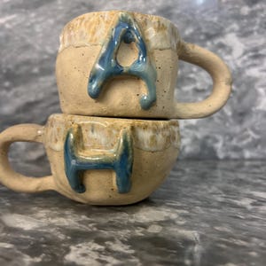 Könnte beinhalten: Zwei gestapelte Keramikbecher mit Henkeln. Die Becher sind beige mit einem getropften Glanzeffekt an der Oberseite. Der Buchstabe "A" befindet sich auf dem oberen Becher und der Buchstabe "H" auf dem unteren Becher, beide in Blau.