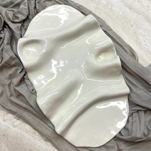 May include: An oval-shaped white ceramic tray with a glossy finish, featuring an irregular, undulating surface with two distinct circular indentations and raised ridges that create a flowing, organic texture across the entire piece.
