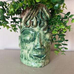 Puede incluir: Una maceta de cerámica verde con forma de rostro con los ojos cerrados y una expresión serena. La maceta tiene una superficie texturizada y está llena de plantas verdes con hojas. El pelo es una mezcla de marrón y verde.