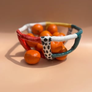 Può includere: Una fruttiera in ceramica colorata piena di arance. Il bordo della ciotola è bianco con sezioni rosse, gialle e verde acqua. Una sezione del bordo è decorata con pois neri. Le arance sono di un arancione brillante.