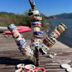 May include: A collection of small, circular ceramic rings featuring various hand-painted patterns in colors including red, blue, yellow, and gold. Several rings are stacked on a wooden branch, while others are scattered on a wooden surface.