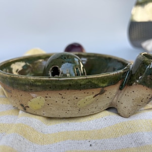 May include: A round ceramic yarn bowl with a speckled beige exterior featuring yellow leaf patterns and a dark green glazed rim. The bowl includes a central dome with a small circular opening and a side notch for yarn, finished with a glossy glaze.