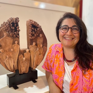 Könnte beinhalten: Eine braune Holzskulptur mit einem halbkreisförmigen Design, das geschnitzte Figuren und strukturierte Details aufweist. Die Skulptur ist auf einem schwarzen Sockel montiert. Der Hintergrund zeigt eine Person mit Brille und einem orange-rosa gemusterten Hemd.