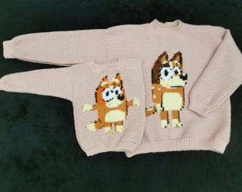 Könnte beinhalten: Zwei rosa gestrickte Pullover mit Comic-Hundebildern. Der größere Pullover hat einen braunen und weißen Hund mit schwarzer Nase und Augen. Der kleinere Pullover hat einen braunen, weißen und orangen Hund mit schwarzen Augen.