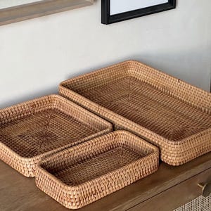 Op de afbeelding: Vier geweven rieten bakken in verschillende maten, gestapeld op een houten oppervlak. De bakken zijn gemaakt van natuurlijke, lichtbruine riet en hebben een rechthoekige vorm.