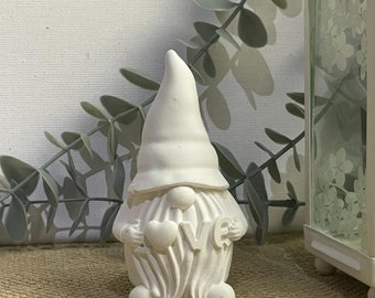 May include: White ceramic gnome figurine holding a heart with the word "LOVE" on it. The gnome is wearing a pointed hat and has a long beard. The gnome is sitting on a burlap surface.