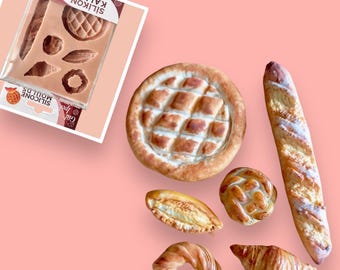 May include: A silicone mold for baking bread, with a variety of shapes, including a round loaf, a baguette, and a crescent roll. The mold is shown with several baked bread items, including a round loaf, a baguette, a crescent roll, a braided bread ring, and a small loaf of bread.