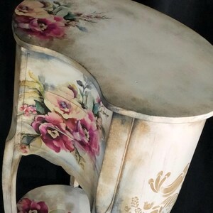 May include: A white painted wooden side table with a curved top and a floral design. The table has a lower shelf and a floral design on the front. The table is distressed and has a vintage look.